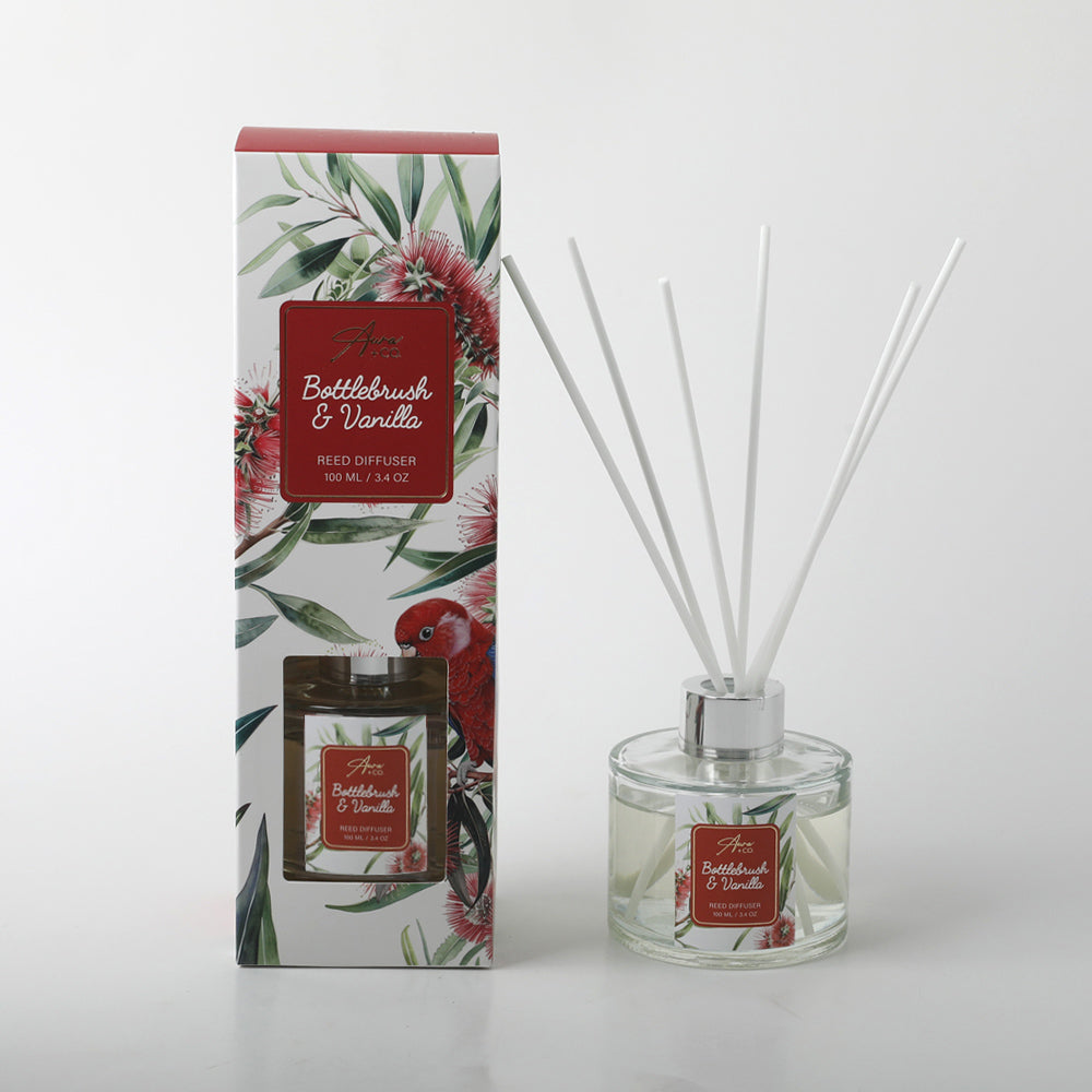 clear round glass bottle with silver collar; white reed sticks; decorative box with bottlebrush flower and red parrot artwork; red label with white text on bottle and box;
