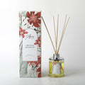 clear glass bottle with silver-coloured cap; yellow liquid inside bottle; natural reed sticks inserted in bottle; packaging with floral print in red and green;