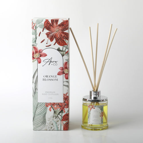 clear glass bottle with silver-coloured cap; yellow liquid inside bottle; natural reed sticks inserted in bottle; packaging with floral print in red and green;