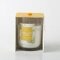 white candle in clear glass holder; yellow and white floral label with golden wattle illustration; packaged in white folded box with yellow top; yellow ribbon pull tab on top of box;