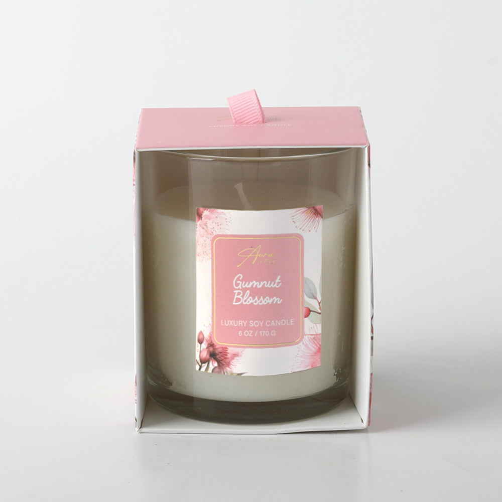 white candle in clear glass container; pink floral label with gumnut blossom illustration; pink cardboard box with front window; pink fabric ribbon pull tab on top;