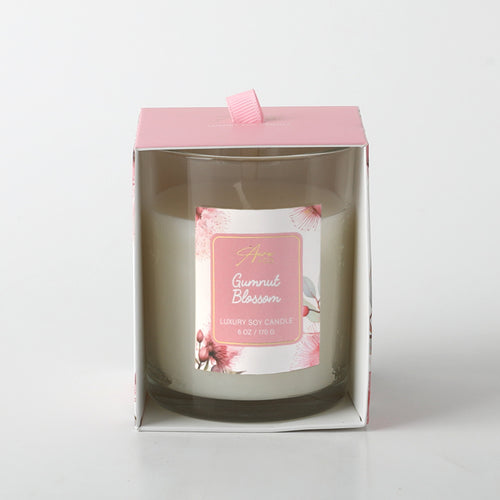 white candle in clear glass container; pink floral label with gumnut blossom illustration; pink cardboard box with front window; pink fabric ribbon pull tab on top;