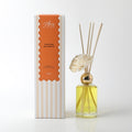 Clear glass diffuser bottle with yellow liquid; Gold-toned collar around bottle neck; Natural coloured reed sticks inserted; Cream-coloured foam flower attached; Product box with orange and beige stripes;