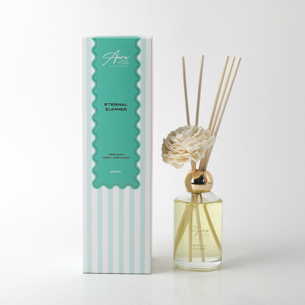 clear glass bottle with light yellow liquid; decorative foam flower attached at top; gold metallic collar around bottle neck; natural coloured reed sticks; packaging box with teal and white stripes;