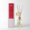 Clear glass bottle with gold cap; Light cream foam flower attached; Natural reed sticks inside bottle; Striped pink and white packaging with red label; Gold printed branding on bottle;