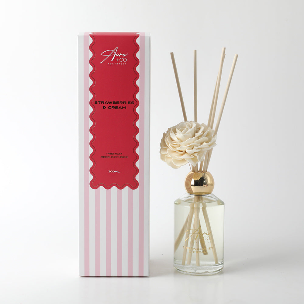 Clear glass bottle with gold cap; Light cream foam flower attached; Natural reed sticks inside bottle; Striped pink and white packaging with red label; Gold printed branding on bottle;