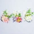 Three carded accessories shown side by side; Artificial flowers in pastel pink, purple and white tones; Green foliage and small buds mixed with blooms; Elastic hair tie base visible behind floral arrangement;