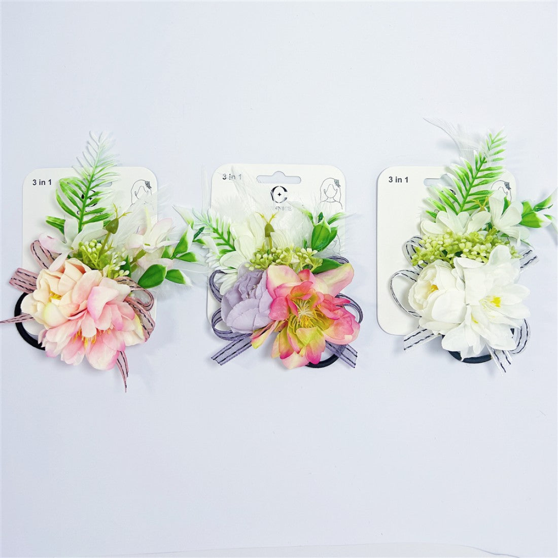 Three carded accessories shown side by side; Artificial flowers in pastel pink, purple and white tones; Green foliage and small buds mixed with blooms; Elastic hair tie base visible behind floral arrangement;