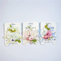 Artificial flower clusters in pastel pinks and whites; Green foliage accents around the blooms; Soft ribbon tails in pale yellow, cream and light blue; Each accessory mounted on white branded backing card;