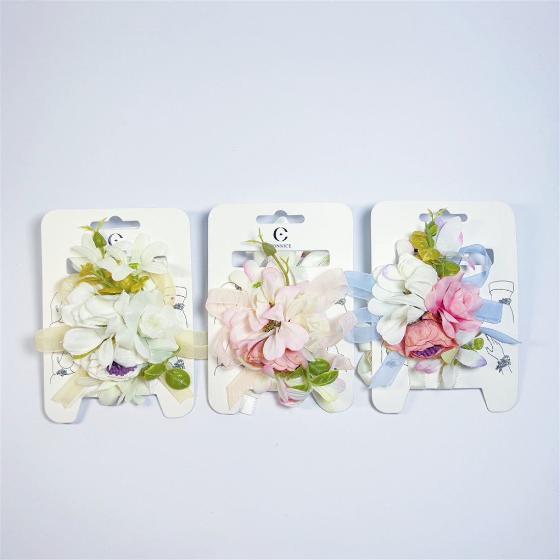 Artificial flower clusters in pastel pinks and whites; Green foliage accents around the blooms; Soft ribbon tails in pale yellow, cream and light blue; Each accessory mounted on white branded backing card;