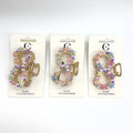 Gold-tone metal bow-shaped claw clip; Decorated with small pastel flower embellishments; Features iridescent rhinestones around the bow; Mounted on white backing card labelled “Hair Accessories”;