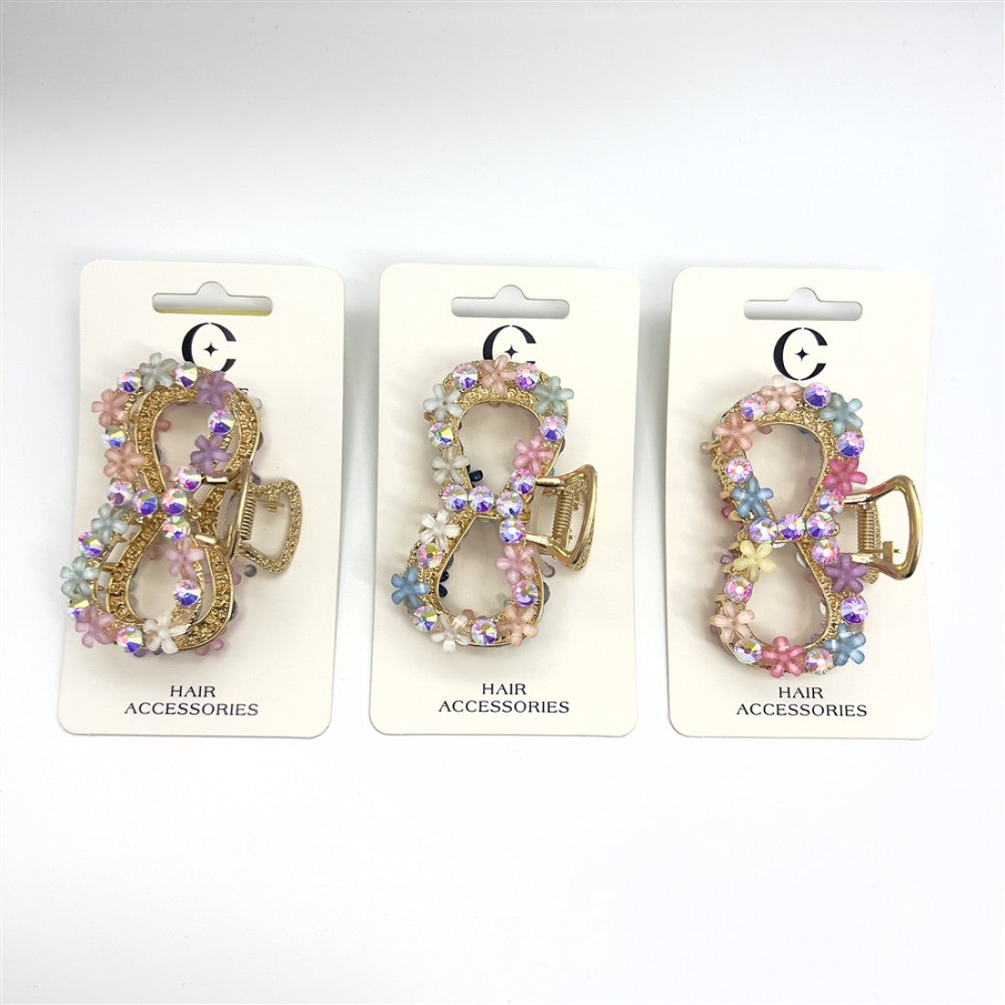 Gold-tone metal bow-shaped claw clip; Decorated with small pastel flower embellishments; Features iridescent rhinestones around the bow; Mounted on white backing card labelled “Hair Accessories”;