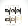 Cross-shaped claw clip design; Approx. 8cm medium size; Made from glossy plastic/PC material; Packaging shows assorted neutral colours including black, brown and clear; Mounted on white backing card with simple branding;
