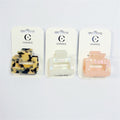 Three square-shaped hair claw clips displayed on branded backing cards; Colours include tortoiseshell, pearly white and soft peach; Glossy acrylic material with cut-out centre rectangle; Metal hinge visible on the side of each claw;
