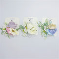 Artificial flower cluster with multiple blooms; Pastel colours including purple, pink, cream and blue tones; Green leaf and foliage accents; Mounted on branded backing card; Metal hair clip backing visible behind flowers;