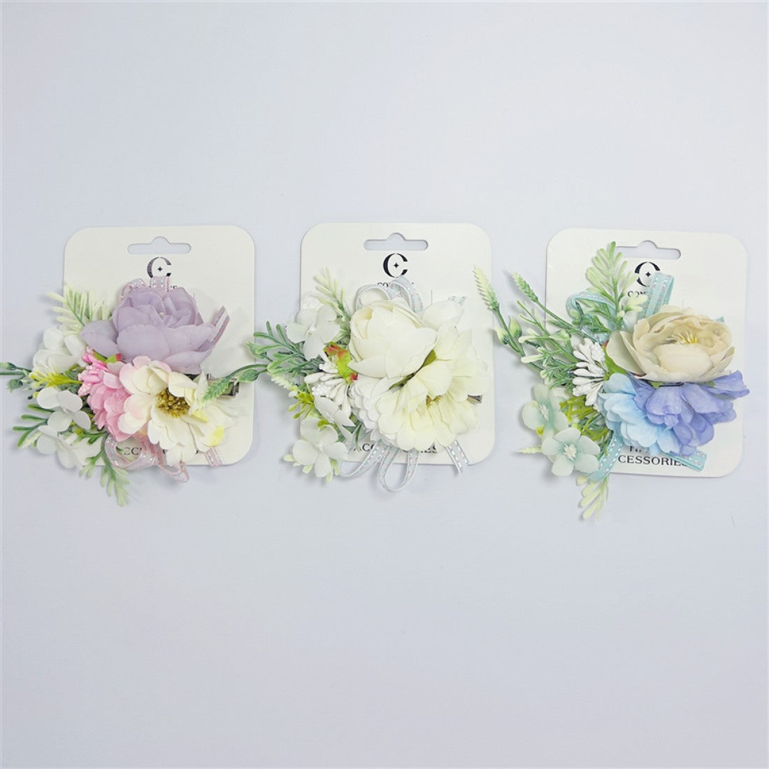 Artificial flower cluster with multiple blooms; Pastel colours including purple, pink, cream and blue tones; Green leaf and foliage accents; Mounted on branded backing card; Metal hair clip backing visible behind flowers;