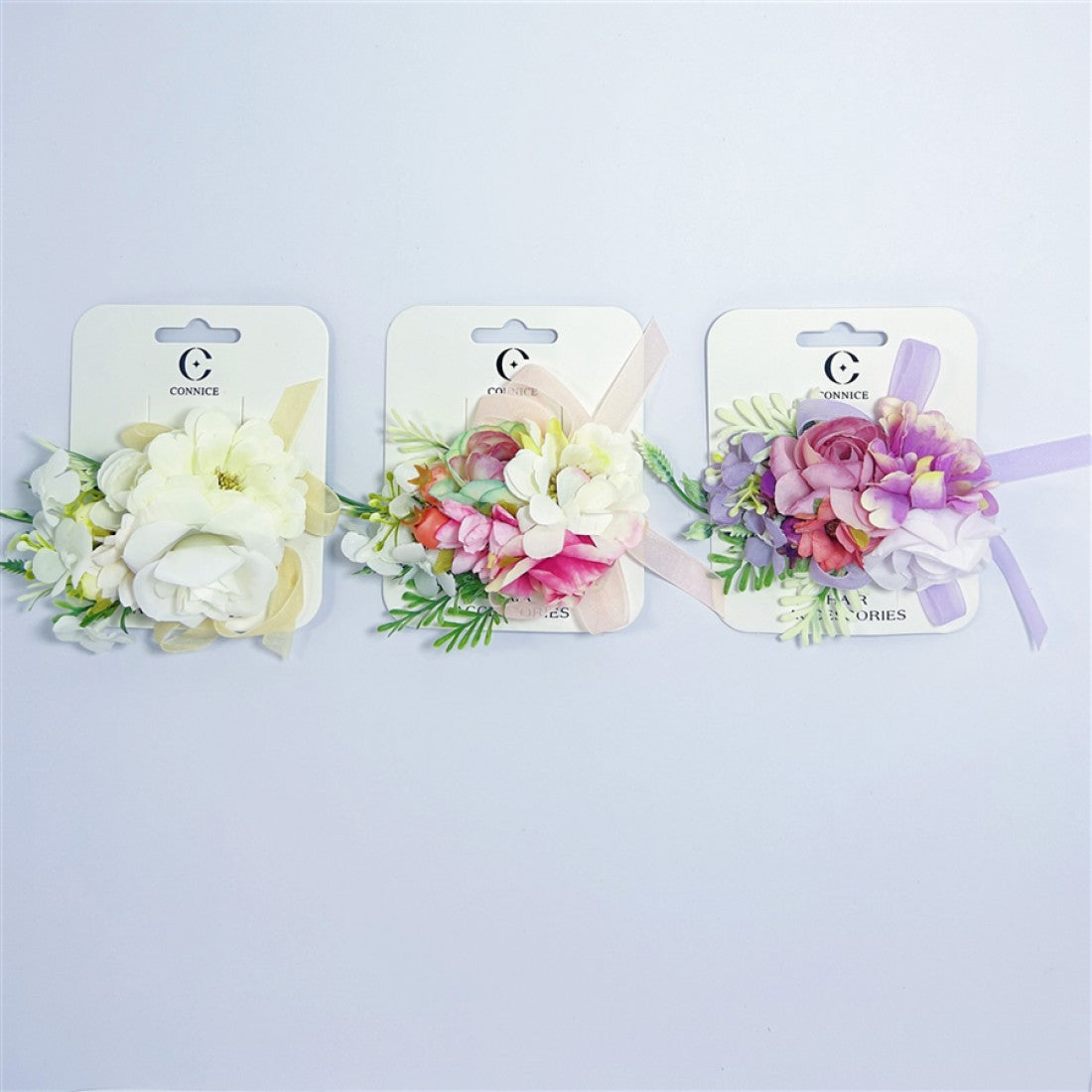 Artificial flower cluster design with layered petals; Available in light, pastel and multicolour arrangements; Includes green leaf accents and small buds; Mounted on white backing card branded “Connice”; Ribbon detail in matching pastel shade on each clip;
