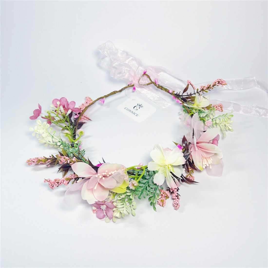 Wreath style floral crown with circular shape; Mixed pastel flowers in pink, cream and yellow tones; Green foliage and small berry-like accents; Pink sheer ribbon tie at the back; Attached branded swing tag on the wreath;