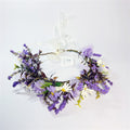 Purple and white artificial flowers arranged in a circle; Green and dark reddish foliage throughout the wreath; Translucent white ribbon tie at the back; Small clustered purple buds and daisy-style blooms;