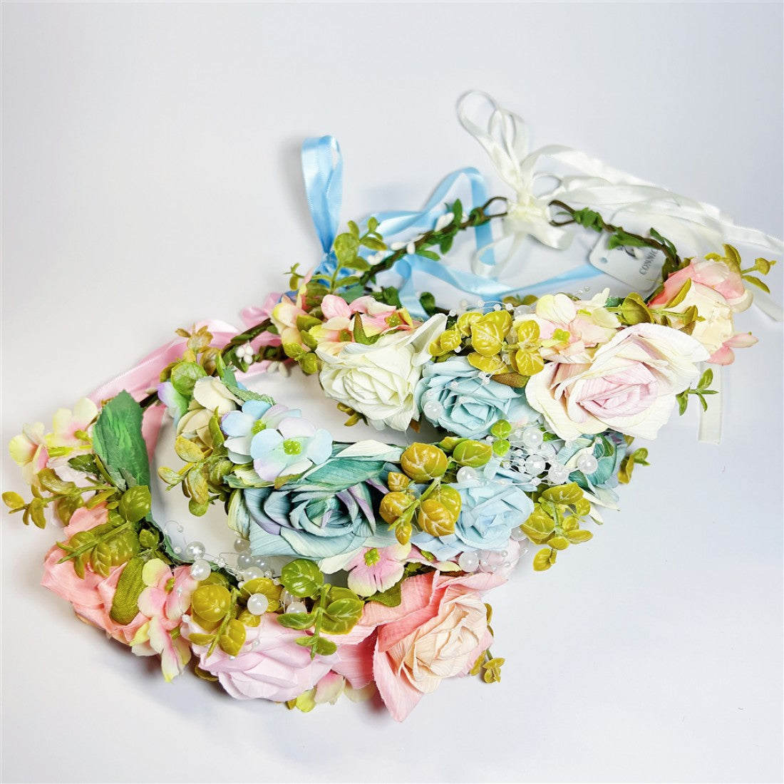 Pastel-coloured artificial roses and small flowers; Green leafy foliage around the wreath; Ribbon ties in soft colours including blue and white; Round floral crown or wreath shape; Small pearl-like bead accents among the flowers;