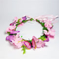 Pink artificial flowers arranged in a circular wreath; Multiple purple and pink butterfly decorations attached; Green faux leaves and buds along the wreath; Light pink ribbon ties at the back; Brown vine-style wreath base;