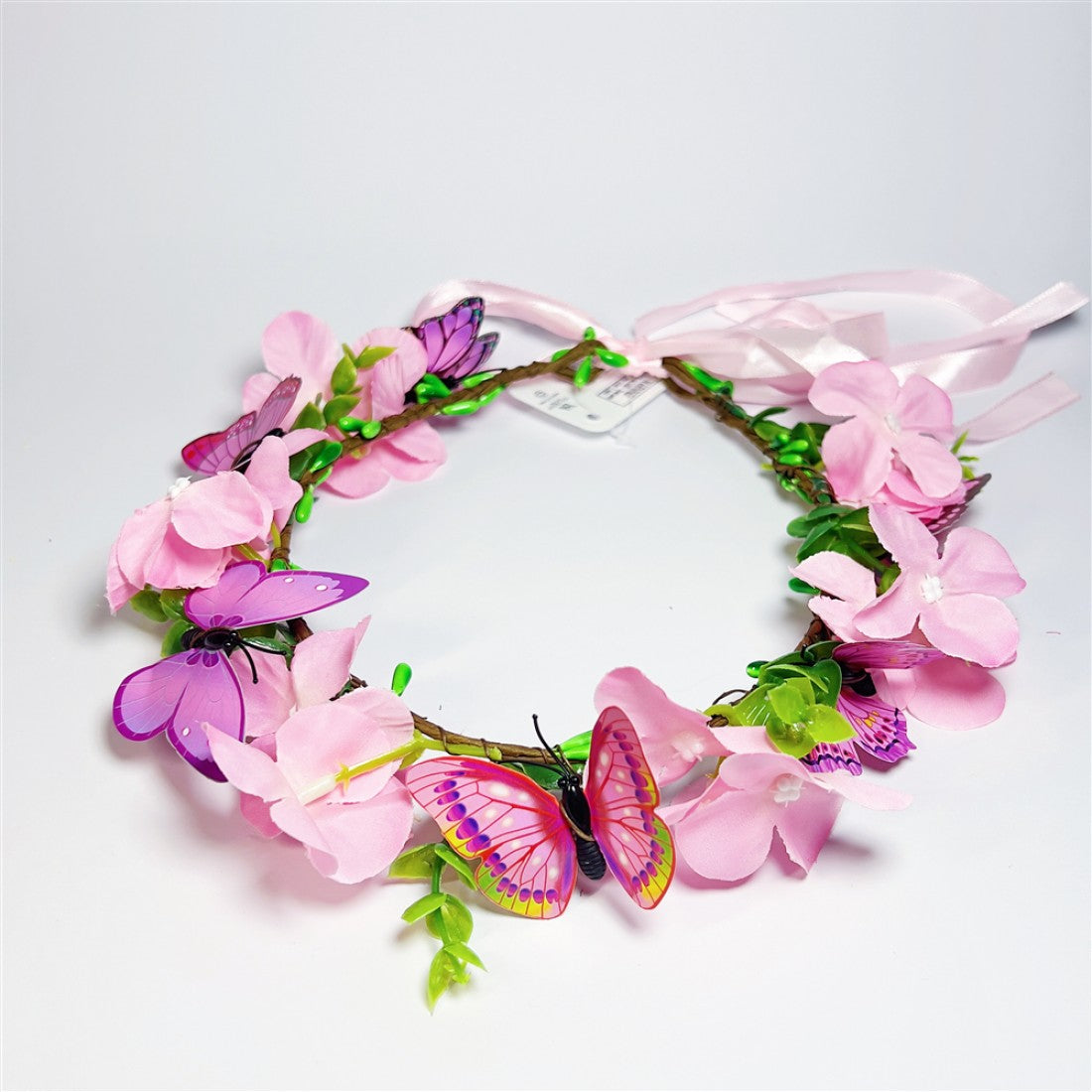 Pink artificial flowers arranged in a circular wreath; Multiple purple and pink butterfly decorations attached; Green faux leaves and buds along the wreath; Light pink ribbon ties at the back; Brown vine-style wreath base;