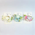 Artificial flower wrist corsages on branded backing cards; Three colour options shown: white, blue, and pink pastels; Mix of small blossoms and larger bloom at centre; Green foliage accents around flowers; Rope-style band attached to each corsage;