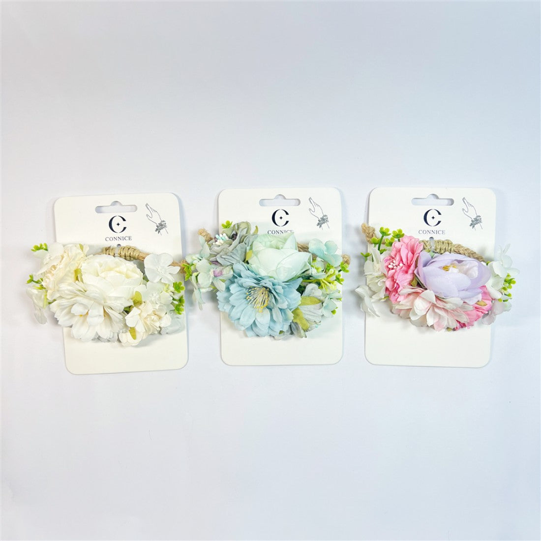 Artificial flower wrist corsages on branded backing cards; Three colour options shown: white, blue, and pink pastels; Mix of small blossoms and larger bloom at centre; Green foliage accents around flowers; Rope-style band attached to each corsage;