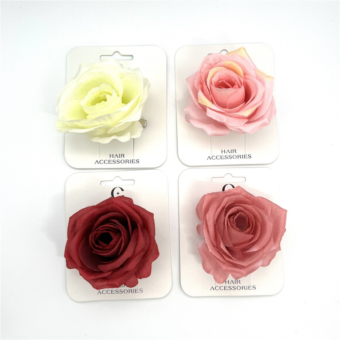 Large rose-shaped fabric flower; Shown in cream, light pink, dusty pink and deep red colours; Mounted on white backing cards labelled “Hair Accessories”; Metal clip visible behind one flower;