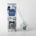 Glass diffuser bottle with silver neck collar; Clear liquid inside bottle; White reed sticks inserted in bottle; Box packaging with blue floral design and bird illustration; Blue label on bottle and box with white text;
