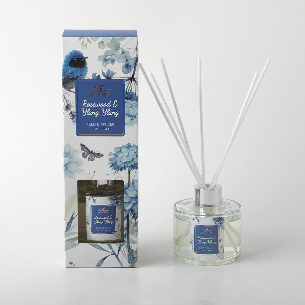 Glass diffuser bottle with silver neck collar; Clear liquid inside bottle; White reed sticks inserted in bottle; Box packaging with blue floral design and bird illustration; Blue label on bottle and box with white text;