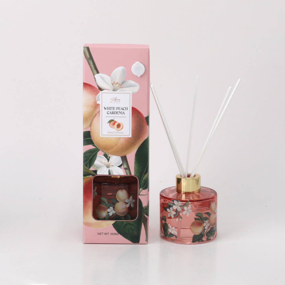 Pink floral decorative cardboard packaging; Clear round glass bottle with peach and flower illustrations; Gold-coloured bottle neck collar; White reed sticks inserted in bottle;