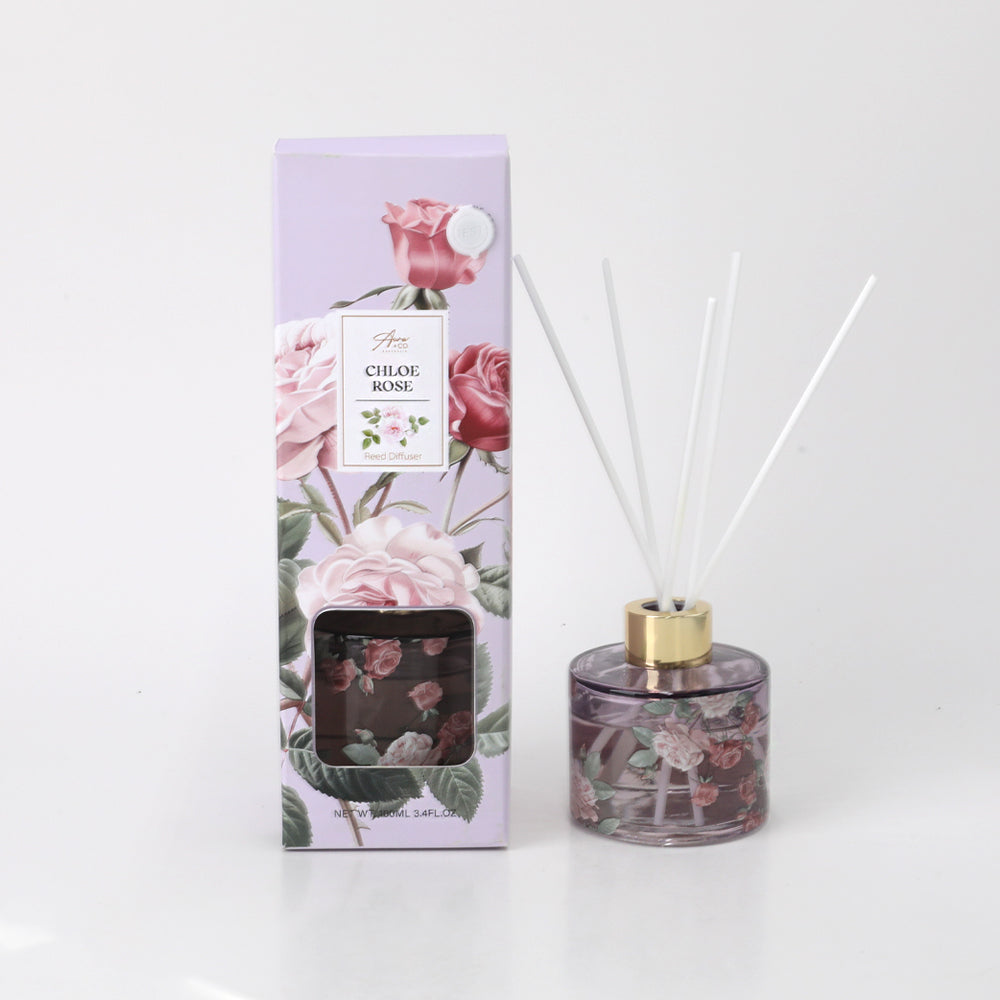 100ml clear round glass diffuser bottle with floral print; gold-coloured neck collar; five white diffuser reeds; packaging box with purple background and rose illustrations;