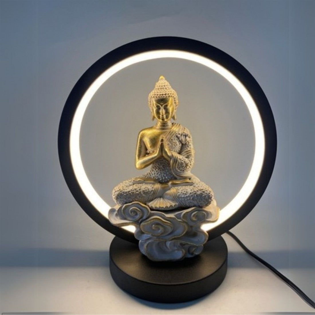 Circular ring light surrounding a central figure; Gold-toned Buddha statue seated in a meditative pose; Cloud-like sculpted base in light grey tones; Black round base with attached power cable;