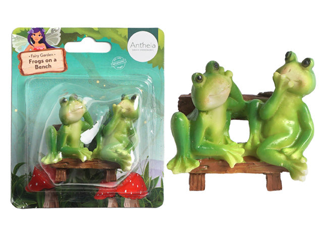 Two green frogs sitting on a brown bench; Bench has a wood-like texture; Figurines are made of resin or plastic; Packaging includes a cardboard backing with fairy garden theme;
