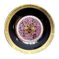 gold-tone circular plate; black inner rim; central Aboriginal artwork with pink and multiple colours; repeated AUSTRALIA text on gold rim;