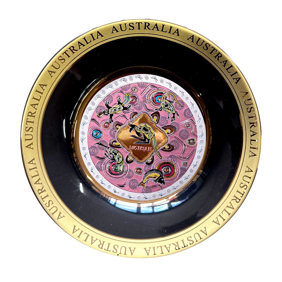 gold-tone circular plate; black inner rim; central Aboriginal artwork with pink and multiple colours; repeated AUSTRALIA text on gold rim;