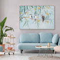 Hand painted canvas; Features three kookaburras on a branch; Light blue and green background; Yellow flowers accents; Framed canvas with slim border;
