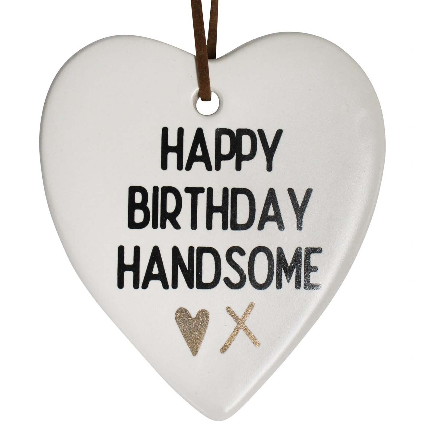 White ceramic heart shape; Black text 'HAPPY BIRTHDAY HANDSOME'; Gold heart and cross design; Brown hanging strap; Smooth matte finish;