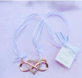 gold-coloured metal pendant; heart outline with rhinestones; infinity symbol integrated; white ribbon tied in a bow; delicate and small size;