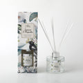 Clear glass round bottle with silver collar; White reed sticks; Floral and bird printed label on bottle and packaging; Tall rectangular packaging with botanical design;