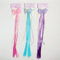 Three long braided hair extensions in pink, blue and purple; Each extension features a large butterfly-shaped bow at the top; Braids are mixed with loose straight strands; Product is displayed on pastel purple backing cards with hooks;