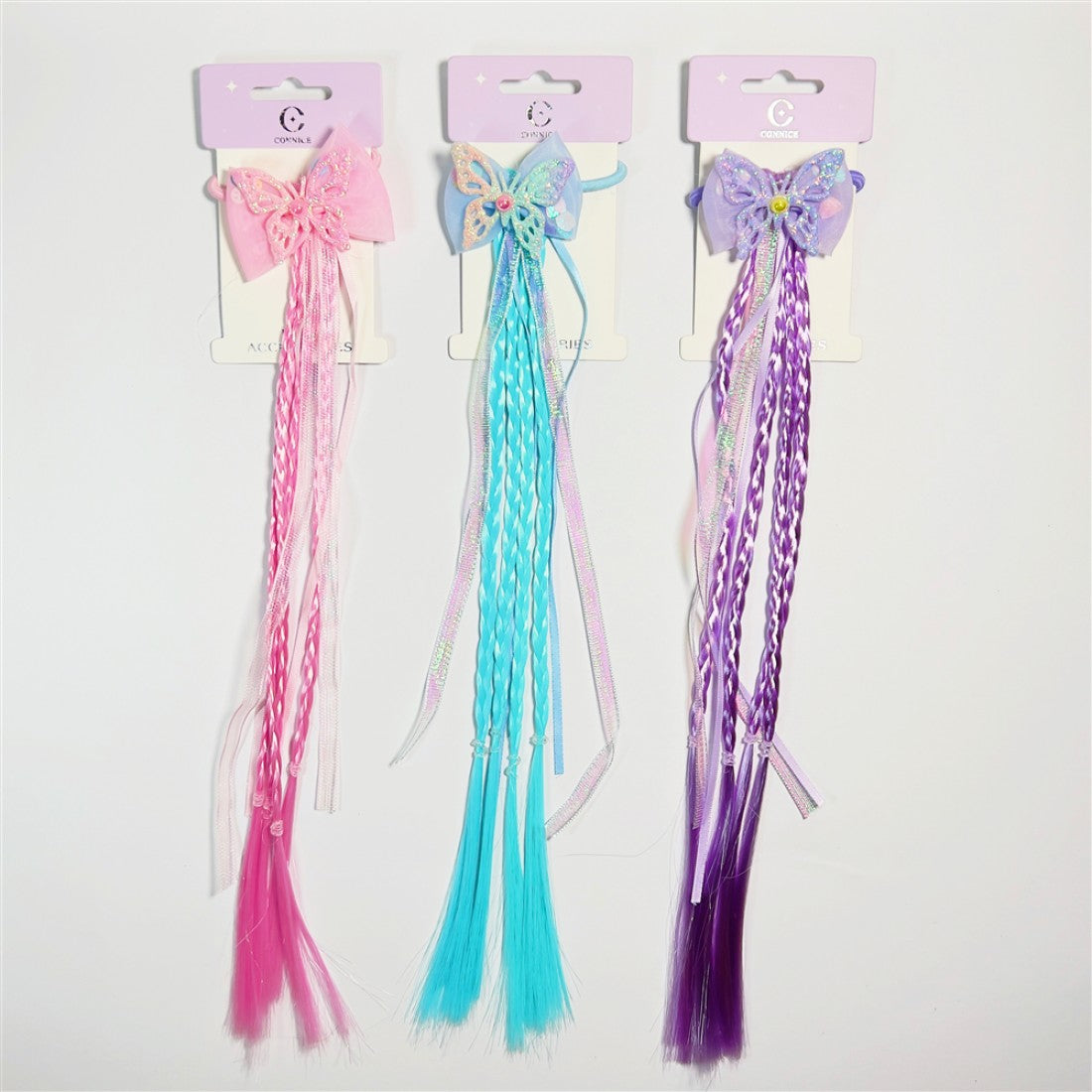 Three long braided hair extensions in pink, blue and purple; Each extension features a large butterfly-shaped bow at the top; Braids are mixed with loose straight strands; Product is displayed on pastel purple backing cards with hooks;