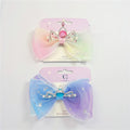 Large layered organza bow in gradient pastel colours; Metallic crown decoration with rhinestones and faux pearls; Glitter accents scattered across bow fabric; Mounted on branded backing card labelled “Kids Accessories”;