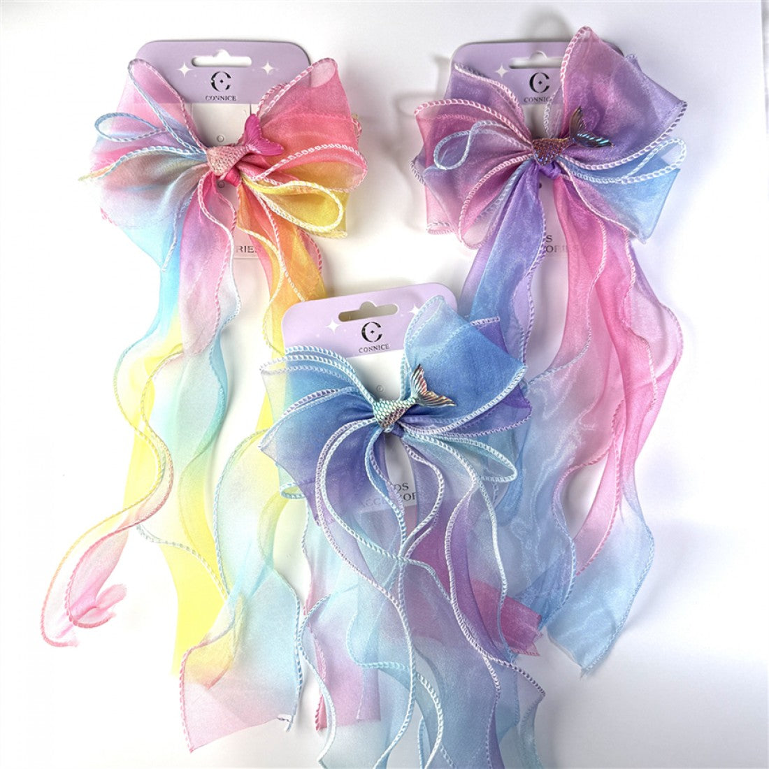 Large organza bow with long flowing ribbon tails; Soft pastel gradient colours including pink, blue, yellow and purple; Small metallic-style mermaid tail charm at the centre of the bow; Packaged on a pale purple backing card with hanging slot;