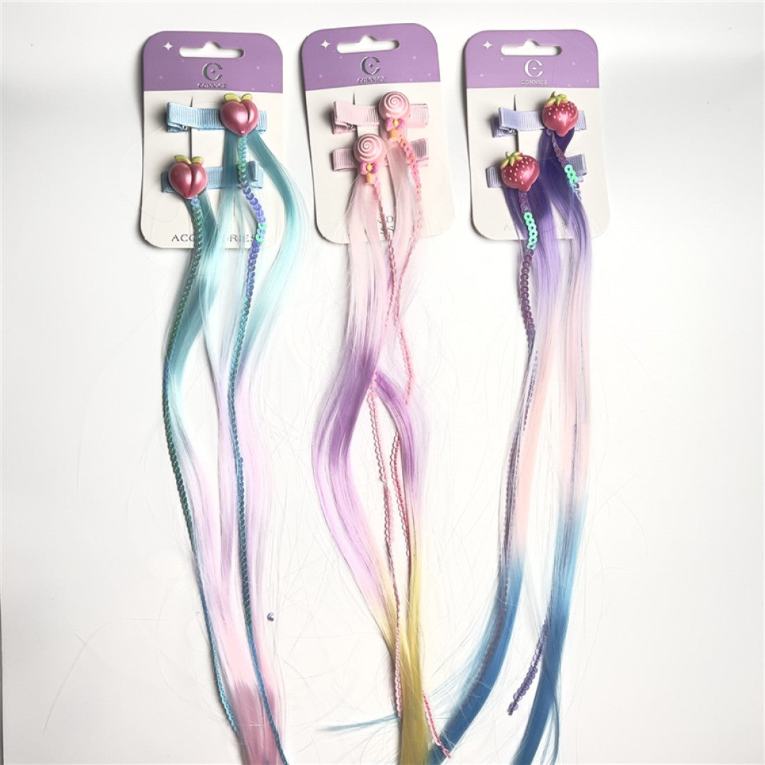 Clip-in hair extensions on backing cards; Rainbow pastel colours including blue, pink, purple and yellow; Fruit-shaped charms such as peach and strawberry; Braided strands mixed with straight synthetic hair;