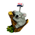grey koala figurine; brown textured tree branch; spring holding a small white hat; hat with red AUS lettering;