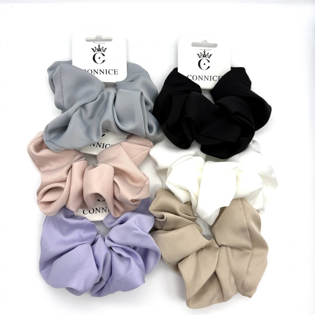 Large fabric scrunchies with a soft satin-like sheen; Shown in multiple colours including black, white, grey, beige and pastels; Attached to branded backing cards; Fabric appears smooth and slightly glossy;