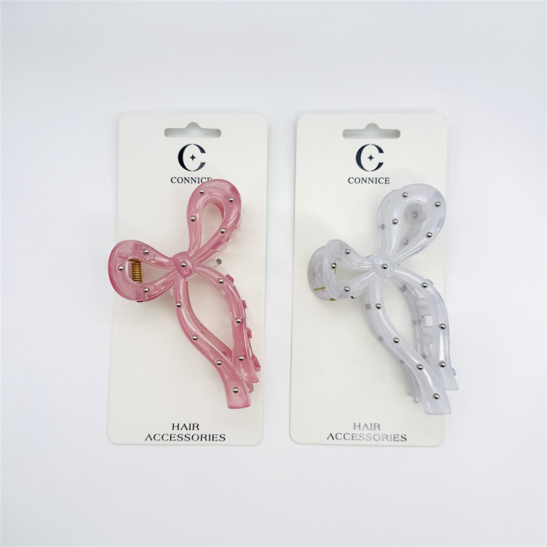 Two bow-shaped claw clips displayed on backing cards; One clip is translucent pink and one is translucent white; Both clips feature small silver bead studs; Gold-toned metal claw mechanism is visible; Packaging is branded "Connice" with "Hair Accessories" text;