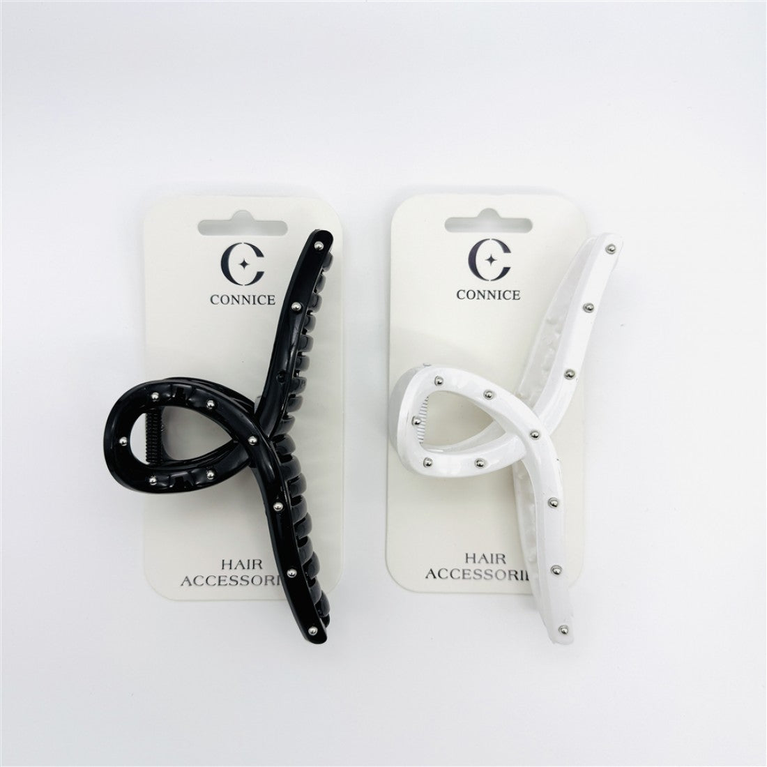 Long cross-shaped claw clip design; One black clip and one white clip shown; Silver bead studs along the arms of the clip; Mounted on branded backing cards labelled "Connice"; Curved teeth visible on inner edge of clip;